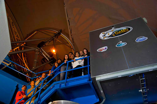 Image of the Gemini South laser propagation team. The team is standing under a large telescope dome with a laser pointed towards it. A metal enclosure containing the laser equipment is visible to the right of the team. Team members are identified from left to right in the caption.