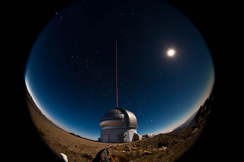 A view of the Gemini South telescope on a night with a bright gibbous moon. A laser beam shoots up into the sky from the telescope enclosure. This image was taken with a fisheye lens.
